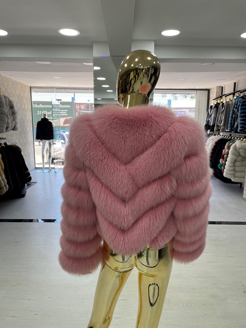May include: A pink fur jacket with a chevron design on the back. The jacket is displayed on a gold mannequin in a clothing store. The sleeves are long and the fur appears soft and plush. The jacket is a light shade of pink.