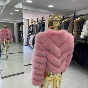 May include: A pink fur jacket displayed on a gold mannequin. The jacket has a chevron design on the back and horizontal stripes on the sleeves. Other fur coats hang in the background. The mannequin is reflected in a large mirror.