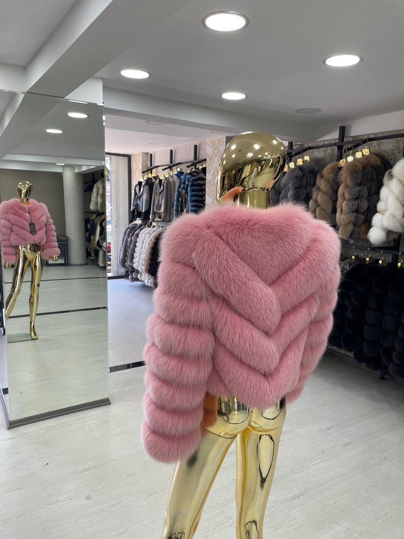 May include: A pink fur jacket displayed on a gold mannequin. The jacket has a chevron design on the back and horizontal stripes on the sleeves. Other fur coats hang in the background. The mannequin is reflected in a large mirror.