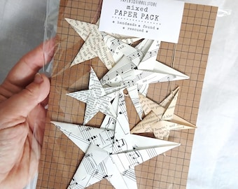 Pode incluir: Um pacote de estrelas de papel feitas à mão em vários tamanhos. As estrelas são feitas de partituras e páginas de texto, com um fundo de grade marrom. O pacote inclui um rótulo que diz "PAPER SQUIRREL STORE mixed PAPER PACK".