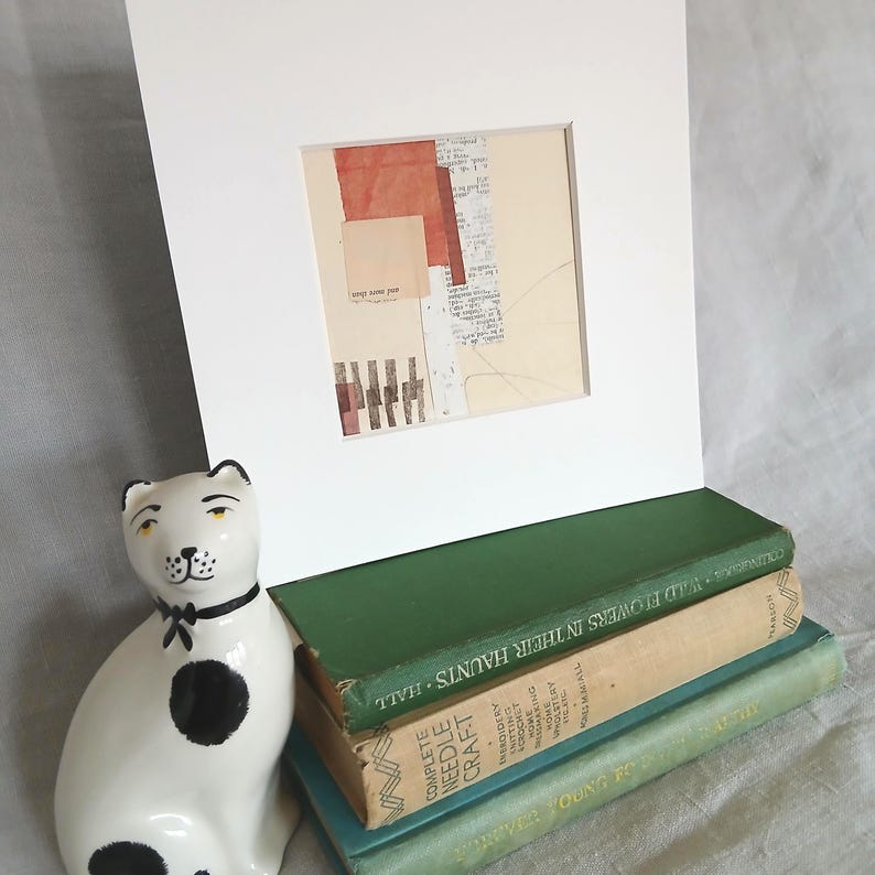 May include: A framed mixed media artwork with a collage of torn paper in shades of red, beige, and white. The artwork is displayed on a white mat, propped up by a stack of vintage books. A ceramic cat figurine with black spots and a bow tie is in the foreground.