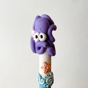 May include: A whimsical, handmade pen topper featuring a purple octopus with large white eyes and a pink flower. The pen is decorated with a starfish and ocean-themed embellishments. The pen is approximately 6 inches long.