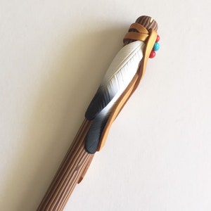 May include: A wooden stick with a brown and black feather attached to it. The feather is wrapped in a tan leather strap with red and turquoise beads.