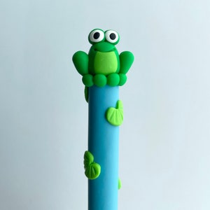 May include: A blue pen with a green frog on top. The frog has big white eyes and a green body with a light green belly. The pen has green leaves on the side.