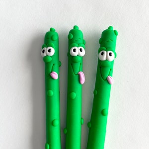 May include: Three green asparagus-shaped pens with cartoon faces. Each pen has a smiling face with large eyes and a pink tongue sticking out. The pens are a bright green color and have small bumps.