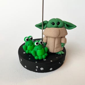 May include: A handmade figurine featuring a green character with large ears and a tan coat, standing next to two green frog figurines. The figures are on a black base with white speckles.