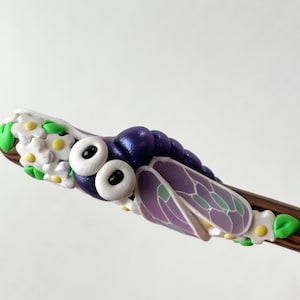 May include: A whimsical, handcrafted dragonfly wand. The wand features a purple dragonfly with white eyes and delicate wings. The wand is decorated with white flowers, green leaves, and yellow accents. The wand is made of polymer clay.