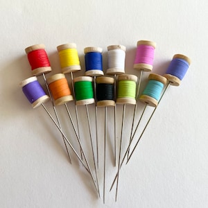 May include: A collection of 12 wooden spools of thread in various colors, including red, orange, yellow, green, blue, purple, pink, white, and black. The spools are arranged in a semi-circle with their metal needles pointing outwards.