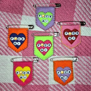 May include: Six felt pennant pins, each featuring a heart shape with white letter beads spelling FUCK AI and a gold star bead. The pins come in various colour combinations including lavender, orange, lime green, pink, and bright orange felt.