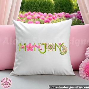 May include: White decorative pillow with the word "Mahjong" embroidered in green, pink, and yellow. The letters are stylized with floral and dragon motifs. The pillow is on a pink pillow with a garden background.