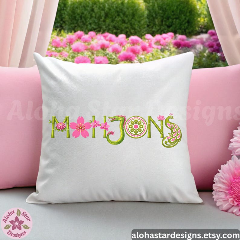 May include: White decorative pillow with the word "Mahjong" embroidered in green, pink, and yellow. The letters are stylized with floral and dragon motifs. The pillow is on a pink pillow with a garden background.
