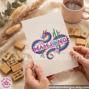 May include: A greeting card featuring the word "MAHJONG" in a decorative font, surrounded by a colorful dragon design. The card is held in hands, with Mahjong tiles and a mug in the background. The card has the text "Aloha Star Designs" and the website address.