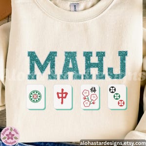 May include: Cream-colored sweatshirt with the word "MAHJ" in teal glitter letters. Below the text are four Mahjong tile graphics. The Aloha Star Designs Etsy shop URL is at the bottom.
