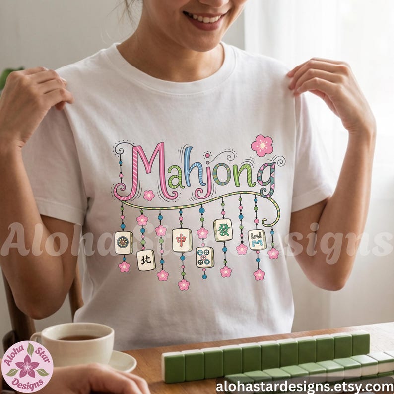 May include: White t-shirt with the word "Mahjong" in colorful script, adorned with floral and tile graphics. The shirt features a playful design with pink and blue accents, perfect for game enthusiasts.