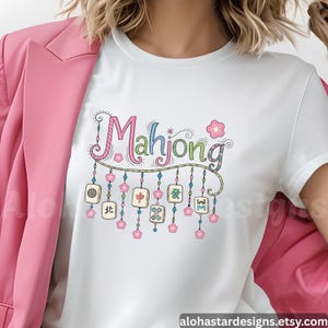 May include: White t-shirt with a colorful "Mahjong" graphic. The design features the word "Mahjong" in a whimsical font, adorned with hanging tiles and floral accents. The shirt is a casual wear item.