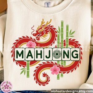 May include: A cream-colored crewneck sweatshirt featuring a graphic print of a red and gold dragon coiled around green bamboo stalks and pink cherry blossoms. The word MAHJONG is printed in dark green letters, with the O replaced by a tile icon.