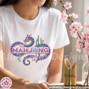 May include: White t-shirt featuring a colorful dragon design with the word "MAHJONG" in a decorative font. The dragon is blue, purple, and pink, with bamboo and flowers. The Etsy shop name is visible.
