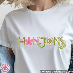 May include: White t-shirt with the word "MAHJONG" in a decorative design. The letters are adorned with pink flowers, green bamboo, and a green dragon. The design includes a peacock and a circular pattern.