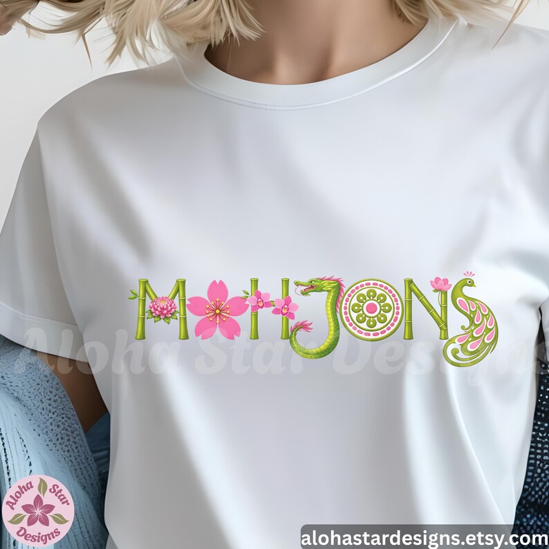 May include: White t-shirt with the word "MAHJONG" in a decorative design. The letters are adorned with pink flowers, green bamboo, and a green dragon. The design includes a peacock and a circular pattern.