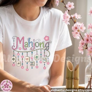 May include: White t-shirt with the word "Mahjong" in colorful, whimsical lettering. Below the word are hanging tiles with Chinese characters and pink flower accents. The design includes the Etsy shop name.