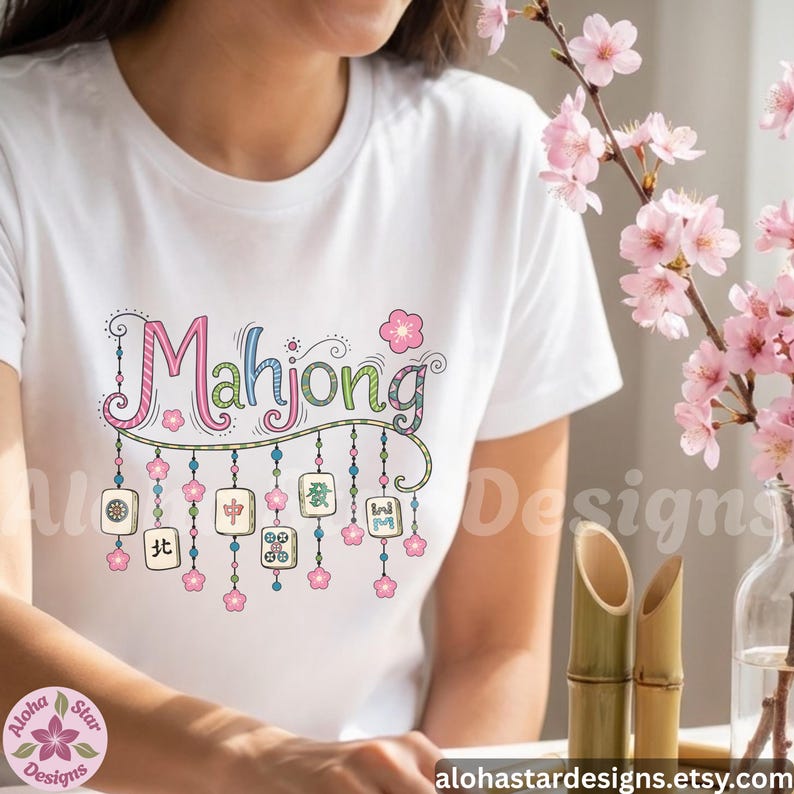 May include: White t-shirt with the word "Mahjong" in colorful, whimsical lettering. Below the word are hanging tiles with Chinese characters and pink flower accents. The design includes the Etsy shop name.