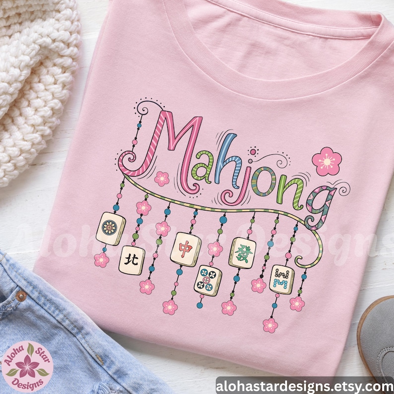 May include: A light pink t-shirt featuring the word "Mahjong" in colorful, whimsical lettering. Below the word are illustrated Mahjong tiles and pink flower accents. The shirt is folded on top of blue jeans.
