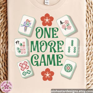 May include: Cream-colored t-shirt featuring a design with Mahjong tiles and the text "ONE MORE GAME" in green. The design includes red flower accents and various Mahjong tile illustrations.