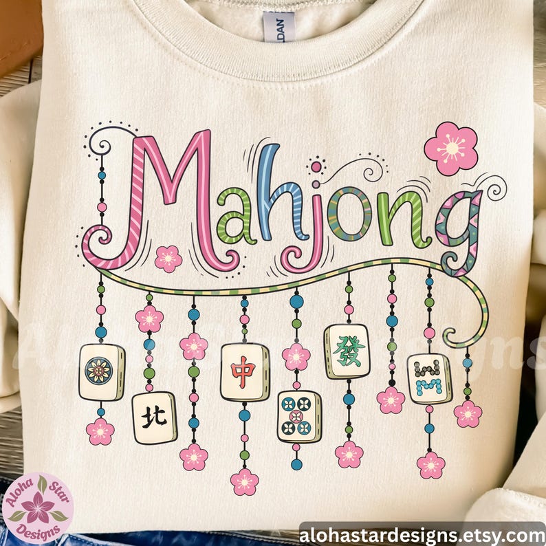 May include: Cream-colored sweatshirt with the word "Mahjong" in colorful, whimsical lettering. The design includes hanging tiles, pink flowers, and decorative beads, evoking a playful, game-themed aesthetic.