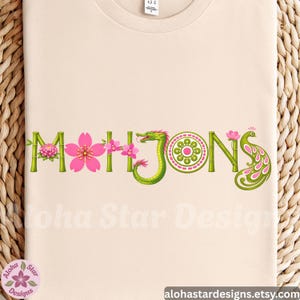 May include: Beige t-shirt with the word "MAHJONG" in green bamboo-style letters, adorned with pink cherry blossoms, a green dragon, and a peacock feather design. The text "Aloha Star Designs" is visible.