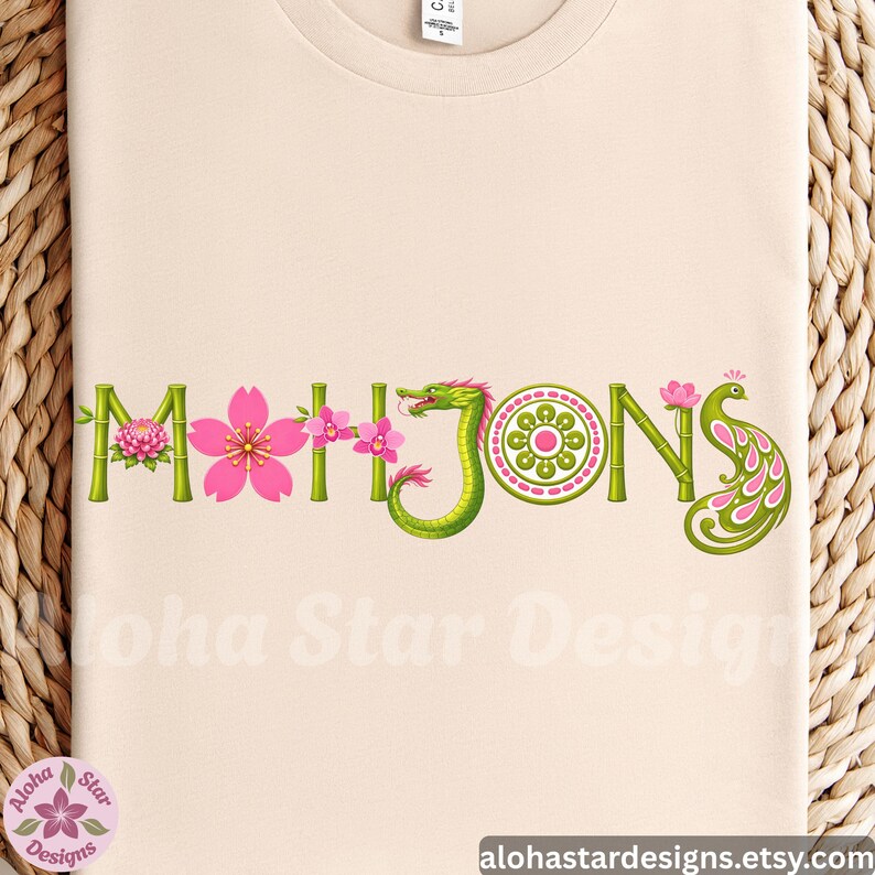 May include: Beige t-shirt with the word "MAHJONG" in green bamboo-style letters, adorned with pink cherry blossoms, a green dragon, and a peacock feather design. The text "Aloha Star Designs" is visible.