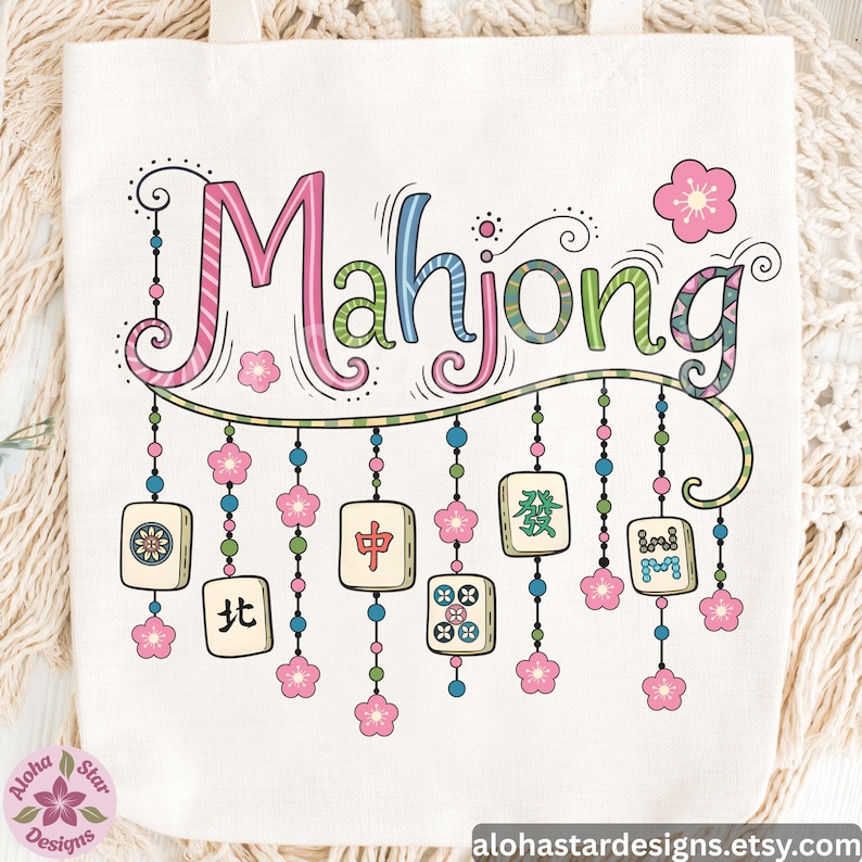 May include: Cream-colored tote bag featuring the word "Mahjong" in colorful, whimsical lettering. Below the word are hanging Mahjong tiles and pink flower accents. The bag has a simple design with a floral logo in the corner.