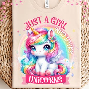 Pode incluir: Uma camiseta bege com um design de unicórnio colorido. O unicórnio tem uma crina e cauda arco-íris, um chifre dourado, e está em um fundo de arco-íris. O texto na camiseta diz "Just a Girl Who Loves Unicorns".