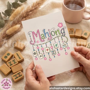 May include: A greeting card with the word "Mahjong" in colorful script, adorned with floral and tile illustrations. The card is held by hands, with wooden Mahjong tiles and a mug of tea in the background. The card's design includes the text "alohastardesigns.etsy.com".