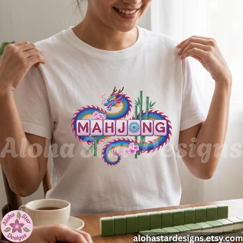 May include: White t-shirt featuring a colorful dragon design with the word "MAHJONG" in a square tile font. The design includes bamboo stalks and pink flowers. A cup of tea and mahjong tiles are visible.