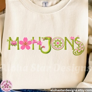 May include: Cream-colored sweatshirt with the word "Mahjong" in a decorative design. The letters are green bamboo with pink flowers and a green dragon. The letter "O" is a green circle with a pink flower design, and the letter "S" is a pink and green peacock.