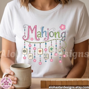 May include: White t-shirt featuring the word "Mahjong" in colorful, whimsical lettering. Below the word are hanging decorative elements, including pink flowers and game tiles. The design is playful and eye-catching, perfect for game enthusiasts.