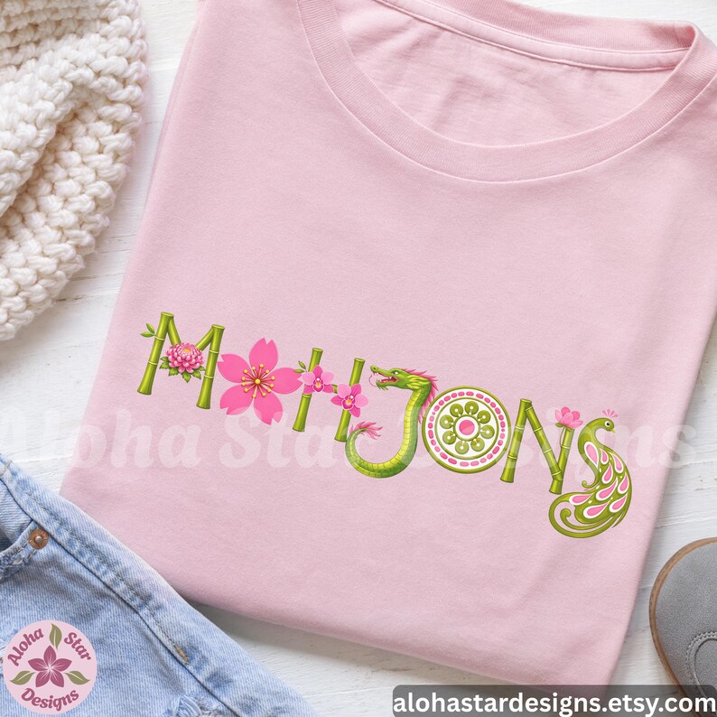 May include: A light pink t-shirt with the word "Mahjong" embroidered in green bamboo-style letters, adorned with pink cherry blossoms and a green dragon. A peacock design is on the right side of the word.