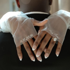 May include: A close-up of a couple's hands with a wedding ring on the woman's finger. The woman's hands are wearing white gloves with the date "1.11.2026" and the text "Mr. & Mrs. Moore" written on them.