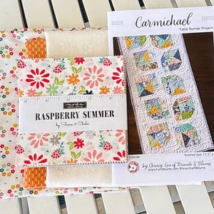 May include: A fabric bundle labeled "Raspberry Summer" with floral patterns, a ruler, and a table runner project pattern. The pattern is titled "Carmichael" and measures 15.5" x 52". The fabric features red, pink, and white flowers.