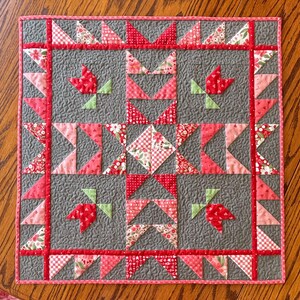 May include: A square, handmade quilt with a gray background and a red and pink floral pattern. The quilt features a central design of triangles, squares, and tulip shapes, framed by a red border. The quilt measures approximately 20 inches by 20 inches.
