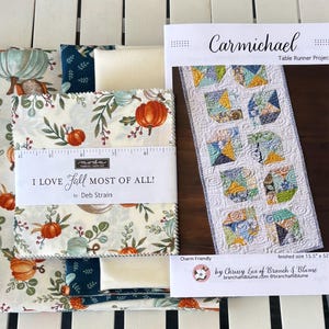 May include: A fabric bundle featuring pumpkin and foliage prints with a paper band labeled I LOVE FALL MOST OF ALL by Deb Strain, alongside a Carmichael Table Runner Project pattern sheet showing a finished size of 15.5 inches by 52 inches.