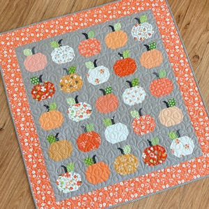 May include: A square quilt featuring a repeating pattern of pumpkin shapes in various orange, white, and gray fabrics. The center panel has a gray background with a swirling quilted texture, surrounded by a wide orange border with white floral prints.