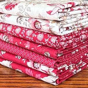 May include: A stack of folded fabric with floral patterns in shades of red, pink, and white. The patterns include large flowers, small dots, and other botanical designs. The fabrics are neatly stacked on a wooden surface.