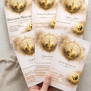 Könnte beinhalten: Ein Satz von sechs Karten mit einem goldenen Lebensbaum-Design, mit deutschem Text. Die Karten haben einen beigen Hintergrund mit goldenen Akzenten. Ein kleiner beiger Beutel mit einem goldenen Lebensbaum-Logo ist ebenfalls sichtbar.