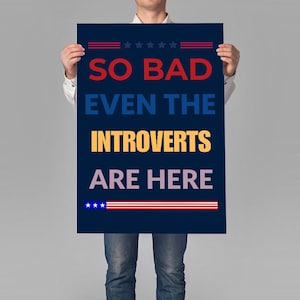 May include: A navy blue poster with the text "SO BAD EVEN THE INTROVERTS ARE HERE" in white and yellow. The poster has red and blue decorative elements, including stars and stripes, and is held by a person.