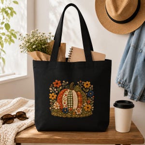 May include: A black tote bag featuring a central graphic of a pumpkin composed of various patterned fabric segments, surrounded by embroidered-style flowers in shades of orange, yellow, and blue, with two sturdy black shoulder straps.
