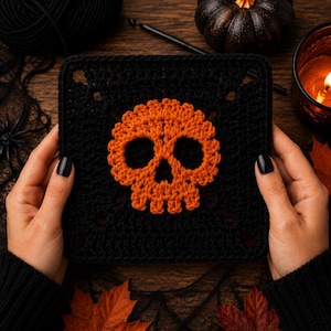 May include: A black crocheted square with an orange skull design. The skull is centered on the square and has detailed features. The square is held by hands with black nail polish. Halloween-themed craft.