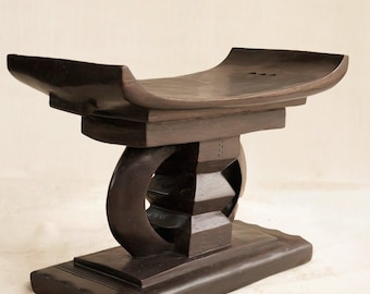 May include: Dark brown wooden stool with a curved seat and a geometric base. The stool has a rectangular base, a central support with a circular cutout, and a seat that curves upwards at the ends. The wood has a polished finish.
