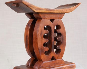 May include: A handcrafted wooden stool with a rich, warm brown finish. The stool features a curved seat and a decorative, openwork design on the central support. The base is rectangular, providing stability. This unique piece is ideal for home decor.