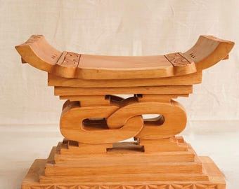 May include: A handcrafted wooden stool with a light brown finish. The stool features a curved seat, decorative carvings, and an interlocking chain design. The base has a geometric pattern. A unique piece of furniture.